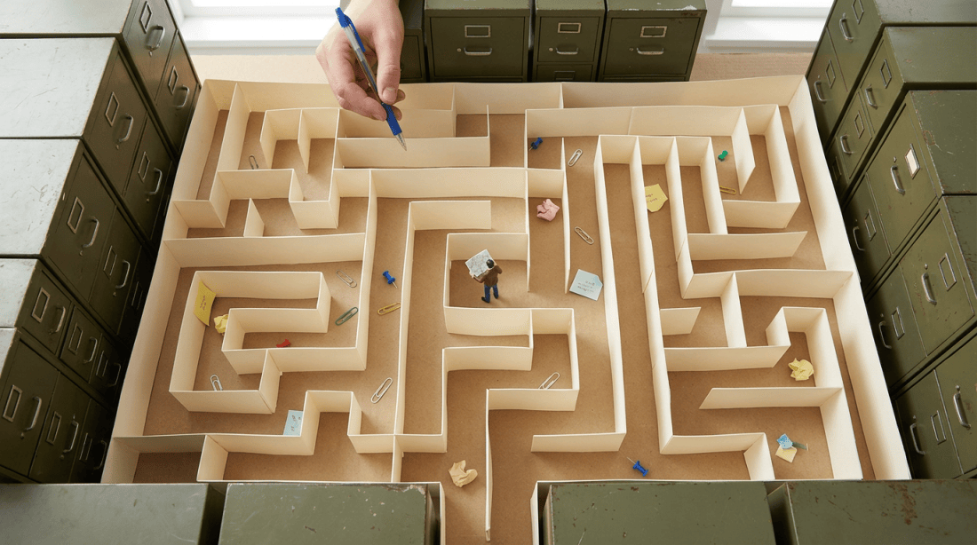 Miniature man standing in a paper maze with notes and paperclips inside an arrangement of filing cabinets